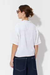 Camisa blanca de algodón con cuello clásico, manga corta y cierre con botones. Presenta bordados de flores, frutas y la palabra 'symphony'.