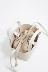 Bolso de mano mini de piel vacuna color blanco roto, con textura granulada. Presenta asas superiores y cordones ajustables laterales. El logo metálico de Bimba y Lola está grabado en el frente.