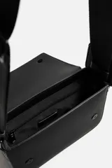 Bandolera negra con acabado engomado y textura acanalada. Tiene un compartimento principal con cierre de solapa y botón a presión, un bolsillo interior con cremallera y un monedero cuadrado en la correa. La correa de hombro es ajustable.
