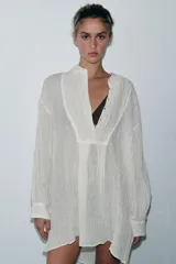 Camisa color crudo de corte holgado, confeccionada en hilatura con lino y tejido con efecto arrugado. Tiene cuello solapa, mangas largas con puños, bajo asimétrico con aberturas laterales y cierre frontal con botones.