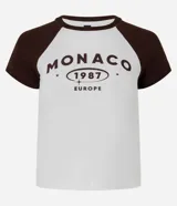 Remera ajustada de algodón con mangas cortas estilo raglán en color marrón oscuro y cuerpo blanco. Presenta un estampado central con el texto "MONACO 1987 EUROPE" en color marrón.