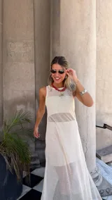 Vestido largo sin mangas, color blanco roto, confeccionado en tejido de red con trama abierta. Se lleva sobre una prenda interior blanca ajustada.