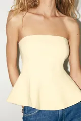 Top blanco de tejido elástico, sin mangas y con escote recto. Tiene corte peplum en la parte inferior y cierre con cremallera oculta en la costura.