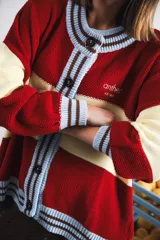 Cardigan tejido a rayas horizontales rojas y crema, con cuello redondo, cierre frontal con botones marrones y mangas largas abullonadas. Presenta ribetes a rayas en cuello, puños y ruedo. Tiene bordado el logo de la marca en el pecho.