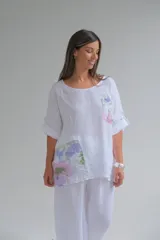 Blusa blanca de lino con cuello redondo y mangas 3/4 con botón. Tiene un bolsillo en el pecho y apliques estampados con motivos florales.