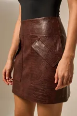 Falda corta de eco-cuero con textura de cocodrilo en tono marrón oscuro. Presenta un diseño de tiro alto con bolsillos frontales superpuestos, uno de ellos con cierre de botón a presión.