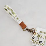 Llavero de muñeca con cinta de tela blanca con patrón de cuadros verde oliva y un estampado de palmera en la parte inferior. Terminado con un detalle de cuero marrón y un gancho metálico de color bronce envejecido.