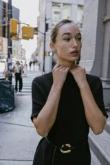 Vestido negro de tejido de punto, corte midi y silueta holgada, con cuello alto acanalado y mangas cortas tipo murciélago. Se ajusta a la cintura con un cinturón fino de efecto piel negro con hebilla dorada en forma de C.