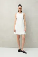 Vestido corto de algodón color verde oliva, de corte recto, con cuello redondo y apliques metálicos en el escote y sisas. Se lleva sobre una camisa negra de manga larga.