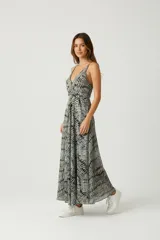 Vestido largo de poliéster con estampado tipo batik o tie-dye en tonos negro, gris y blanco. Presenta escote en V con ribete decorativo, tirantes finos y talle ajustado con nudo o cruce frontal, y falda evasé.