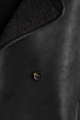 Chaqueta tipo gamulán de color negro con forro de corderito sintético al tono. Presenta solapas anchas, cierre frontal con botones y un diseño entallado al cuerpo.