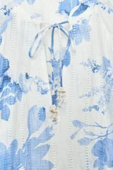 Camisa blanca con estampado de flores azules, cuello redondo con escote en pico y lazo, manga larga con puños elásticos y detalle de tejido perforado.