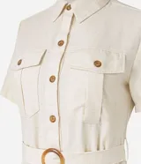 Vestido midi color crema, tipo camisero, con cuello, manga corta y cierre frontal de botones. Presenta bolsillos utilitarios en el pecho y la cadera, y un cinturón ajustable con hebilla redonda de madera. Confeccionado en sarga de algodón.