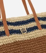 Bolso tipo tote de paja tejida con diseño de rayas horizontales en color natural, marrón y azul marino. Posee asas de hombro en cuero sintético color camel y un dije metálico circular en el frente.