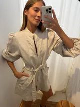 Mono corto de lino color beige claro, con cuello mao, cierre frontal, mangas globo largas y cinto para ajustar a la cintura.