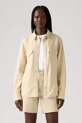 Camisa de manga larga color beige claro, estilo western, con cuello y botones a presión frontales. Presenta dos bolsillos de parche con solapa en el pecho y un pequeño tag rojo en el bolsillo derecho.