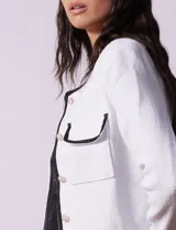 Chaqueta de tweed blanca con ribete negro, escote a la base, abertura central con botones, bolsillos delanteros y puños con aplique de botones perlados.