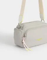 Bandolera gris claro de tela deportiva con bolsillo frontal con cierre y tag de goma. Tiene correa regulable desmontable, acceso mediante cierre superior y bolsillos laterales con cierre y pullers de cordón y goma.