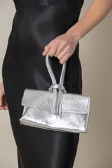 Cartera de mano plateada de cuero laminado con cierre superior y asa doble con nudo. Incluye correa extraíble para colgar.