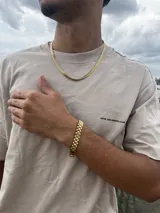 Conjunto de joyería de color dorado, compuesto por una cadena de eslabones tipo cubana o espiga y una pulsera a juego con eslabones anchos y planos. El conjunto se luce sobre una camiseta beige de manga corta con texto estampado en negro.