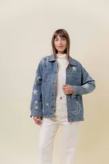 Camisa de denim de lavado medio, con calce relajado y ligeramente oversize. Presenta bordados o estampados de inspiración celestial (soles, lunas y estrellas) en color claro en la parte delantera y mangas. Tiene cuello, bolsillos delanteros con tapa y cierre frontal con botones.