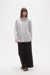 Camisa de algodón blanca de corte holgado, con cuello camisa y manga larga. Presenta un estampado de finas rayas verticales negras y un bolsillo frontal con rayas horizontales.