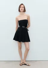 Vestido corto negro, sin mangas, con escote palabra de honor y falda evasé con tablas. Presenta cierre de cremallera en la parte posterior.