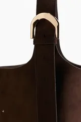 Bolso tipo bombonera de color marrón oscuro, con textura de gamuza o nobuck. Posee un asa de hombro ancha y ajustable con hebilla metálica dorada.