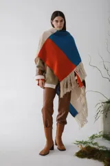 Ruana tejida con diseño geométrico en bloques de color beige, azul y naranja, con flecos en el borde inferior.