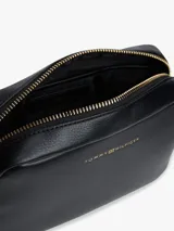 Cartera tipo cámara cruzada (crossbody) color negro, con acabado liso y logo metalizado de Tommy Hilfiger en el frente. Posee un compartimento principal con cierre de cremallera y un asa tejida ajustable.