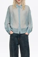 Camisa de corte boxy en color celeste, confeccionada en tejido semitransparente con apliques de pequeñas piedras brillantes. Posee cuello camisero, cierre frontal con botones y mangas largas con puños con sardineta.