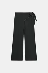 Pantalón negro de tiro medio con cremallera lateral oculta, corte ancho y lazo decorativo en la cadera. Detalle de pespuntes blancos a contraste.