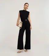 Mono negro sin mangas, con cuello redondo y cierre tipo gota en la espalda. Presenta hombreras marcadas, cintura ajustable con cinturón de lazo y hebilla metálica irregular, y pantalón ancho de pierna acampanada.