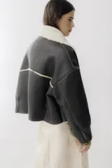 Campera corta de cuero color marrón oscuro con interior de piel sintética color crema. Tiene cuello con solapas, cierre frontal con botones a tono, dos bolsillos delanteros y puños con detalle de piel.
