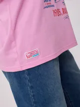 Remera blanca de algodón con estampado frontal de estilo playero con texto "Welcome to the Surf Camp" y dibujo de una casa de playa con palmeras y tablas de surf.