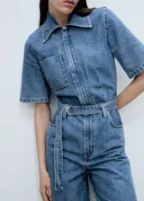 Mono largo de denim azul con cuello camisero, mangas cortas y cierre frontal con cremallera oculta. Incluye cinturón ajustable con hebilla metálica y bolsillo de parche en el pecho.