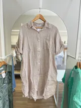 Vestido camisero corto color beige, confeccionado en lino. Tiene cuello camisero, mangas cortas con botón para sujetar, cierre frontal con botones y un bolsillo en el pecho.