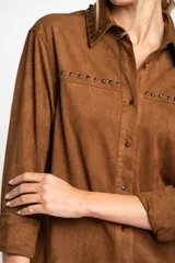Camisa marrón de gamuza vegana con cuello clásico, cierre frontal con botones y mangas largas con puños abotonados. Presenta detalles de tachas metálicas en cuello y charreteras.