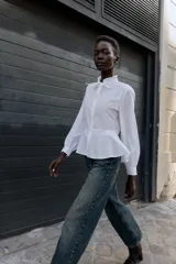 Camisa blanca de popelín con cuello redondo con volante y lazo, manga larga y bajo peplum. Cierre frontal con botones.