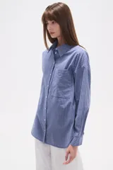 Camisa de algodón blanca de corte holgado, con cuello camisa y manga larga. Presenta un estampado de finas rayas verticales negras y un bolsillo frontal con rayas horizontales.