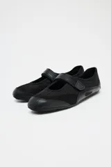 Zapato tipo bailarina color negro, con capellada de cuero sintético y rejilla. Tiene tira con velcro en el empeine y suela plana.