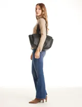 Cartera tote negra de ecocuero con base redondeada y pliegues decorativos. Cuenta con una solapa central con herraje dorado en forma de "T" y asas dobles para hombro y mano. El interior tiene triple compartimento.