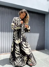 Vestido largo de manga larga, con cuello camisero y estampado de hojas tropicales en blanco y negro. El diseño presenta un corte amplio con volados en la falda.