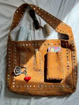 Cartera bandolera marrón de cuero con remaches metálicos plateados. Tiene correa ancha regulable, bolsillo externo y argolla para colgar accesorios.