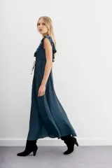 Vestido largo negro sin mangas, con escote redondo, espalda baja y volados en el escote. Ajuste en la cintura con cordón y terminaciones metálicas.