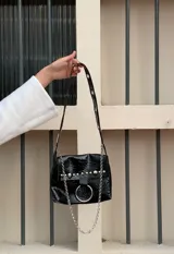Cartera bandolera negra de ecocuero con textura arrugada, tachas plateadas, cadena decorativa y aro metálico.