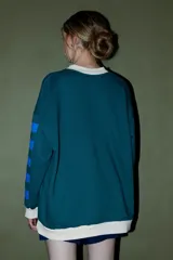 Buzo verde oscuro con cuello redondo y puños y dobladillo en color crema. Presenta un parche ovalado con el texto "CORAL" en el pecho y detalles de franjas azules en las mangas.
