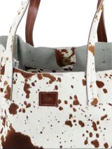 Bolso tote de cuero con pelo, estampado de vaca en blanco y marrón. Tiene asas de cuero marrón y un pequeño parche de cuero marrón con logo grabado en el frente.