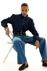 Camisa de denim azul oscuro con cuello mao y volados en cuello y puños. Tiene mangas largas abullonadas con botón en los puños, botonadura frontal y bajo redondeado.