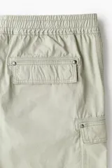 Short cargo color verde claro en popelina de algodón, con cintura elástica ajustable con cordón, bolsillos laterales, bolsillos cargo con solapa y botón a presión en piernas y traseros, y aberturas laterales en el bajo.